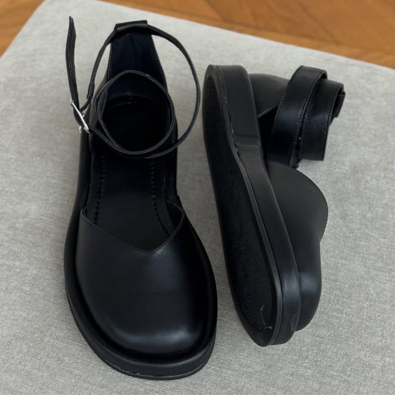 May include: Black leather Mary Jane style shoes with a thick sole. The shoes have a strap that buckles around the ankle and a strap that goes around the foot. The shoes are sitting on a light gray surface.