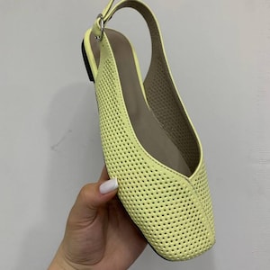 May include: A pale yellow slingback flat shoe with a perforated design. The shoe has a square toe and a thin strap that goes around the heel. The sole is black, and the interior is a light brown color.