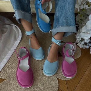 May include: A selection of colorful suede platform sandals in shades of blue and fuchsia. The sandals feature ankle straps with silver buckles and a rounded toe design. One pair is held up by a person wearing blue jeans.