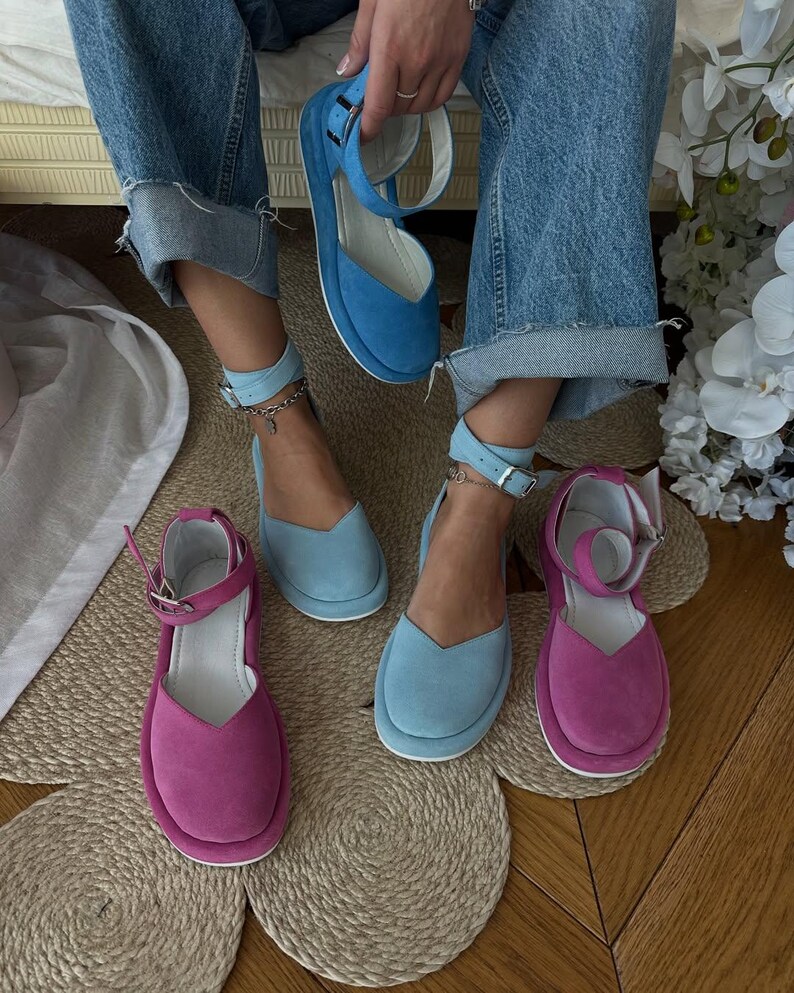 May include: A selection of colorful suede platform sandals in shades of blue and fuchsia. The sandals feature ankle straps with silver buckles and a rounded toe design. One pair is held up by a person wearing blue jeans.
