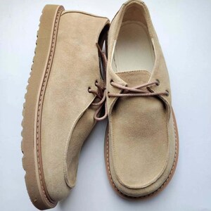 Pu&ograve; includere: Un paio di scarpe in camoscio beige con un design stringato. Le scarpe hanno una suola spessa e strutturata e una punta arrotondata. I lacci e la suola sono di una tonalit&agrave; beige leggermente pi&ugrave; scura. Le scarpe sono esposte su sfondo bianco.