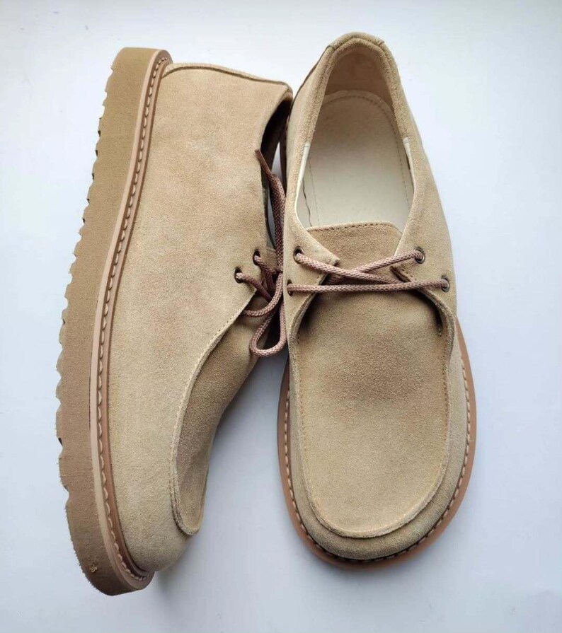 Pu&ograve; includere: Un paio di scarpe in camoscio beige con un design stringato. Le scarpe hanno una suola spessa e strutturata e una punta arrotondata. I lacci e la suola sono di una tonalit&agrave; beige leggermente pi&ugrave; scura. Le scarpe sono esposte su sfondo bianco.