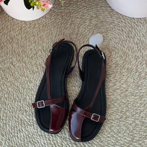 May include: A pair of burgundy leather sandals with black soles. The sandals feature a T-strap design with adjustable buckles for a secure fit. The shoes are displayed on a woven beige surface, with floral arrangements in the background.
