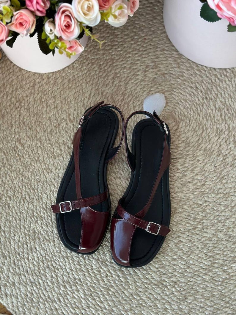 May include: A pair of burgundy leather sandals with black soles. The sandals feature a T-strap design with adjustable buckles for a secure fit. The shoes are displayed on a woven beige surface, with floral arrangements in the background.