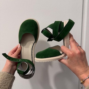 May include: Green suede sandals with a strap and buckle closure. The sandals have a round toe and a cream-colored sole. The strap wraps around the ankle. The shoes are held up by a person's hands.