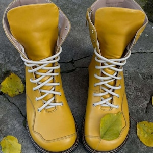 May include: A pair of bright yellow leather boots with white laces. The boots have a rounded toe and a high ankle design. A green leaf rests on the front of one boot. The boots are set on a gray stone surface.