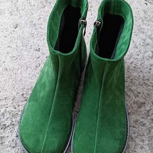 May include: A pair of green suede ankle boots. The boots have a rounded toe, a zipper on the side, and a black sole. The boots are made of a soft, textured material and appear to be unworn.