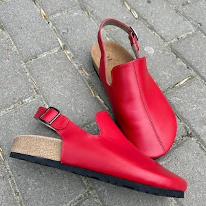 May include: A pair of red leather clogs with a slingback strap and buckle closure. The clogs have a cork footbed and a black sole. The shoes are displayed on a gray brick surface.