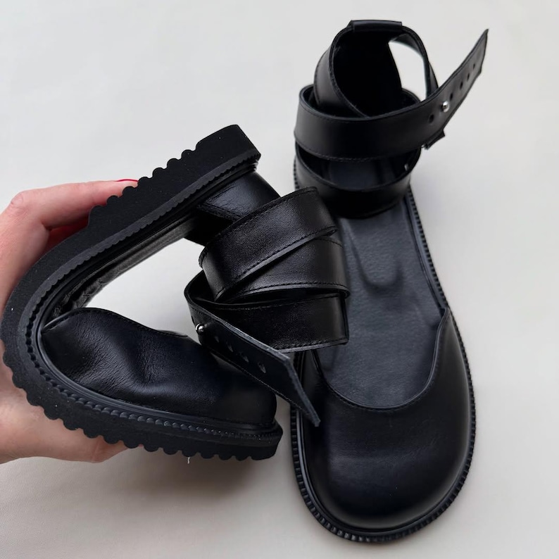 May include: Black leather sandals with a rounded toe and thick, ridged soles. The sandals feature multiple straps that wrap around the ankle and foot, secured with buckles. The design is modern and minimalist.