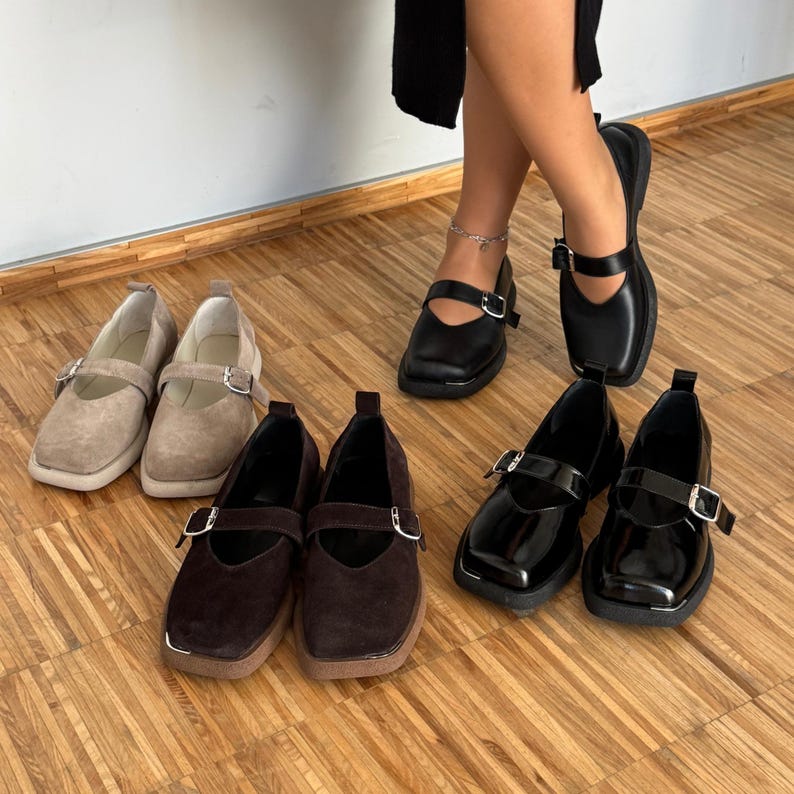 May include: An assortment of Mary Jane style shoes in various colors and materials. The shoes include tan suede, dark brown suede, and black patent leather. Each pair features a strap with a silver buckle and a square toe.
