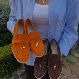 May include: A pair of bright orange suede loafers and a pair of dark brown suede loafers. Each shoe features a horizontal strap across the vamp adorned with a small gold-toned metal padlock and key charm hanging from the center of the strap.