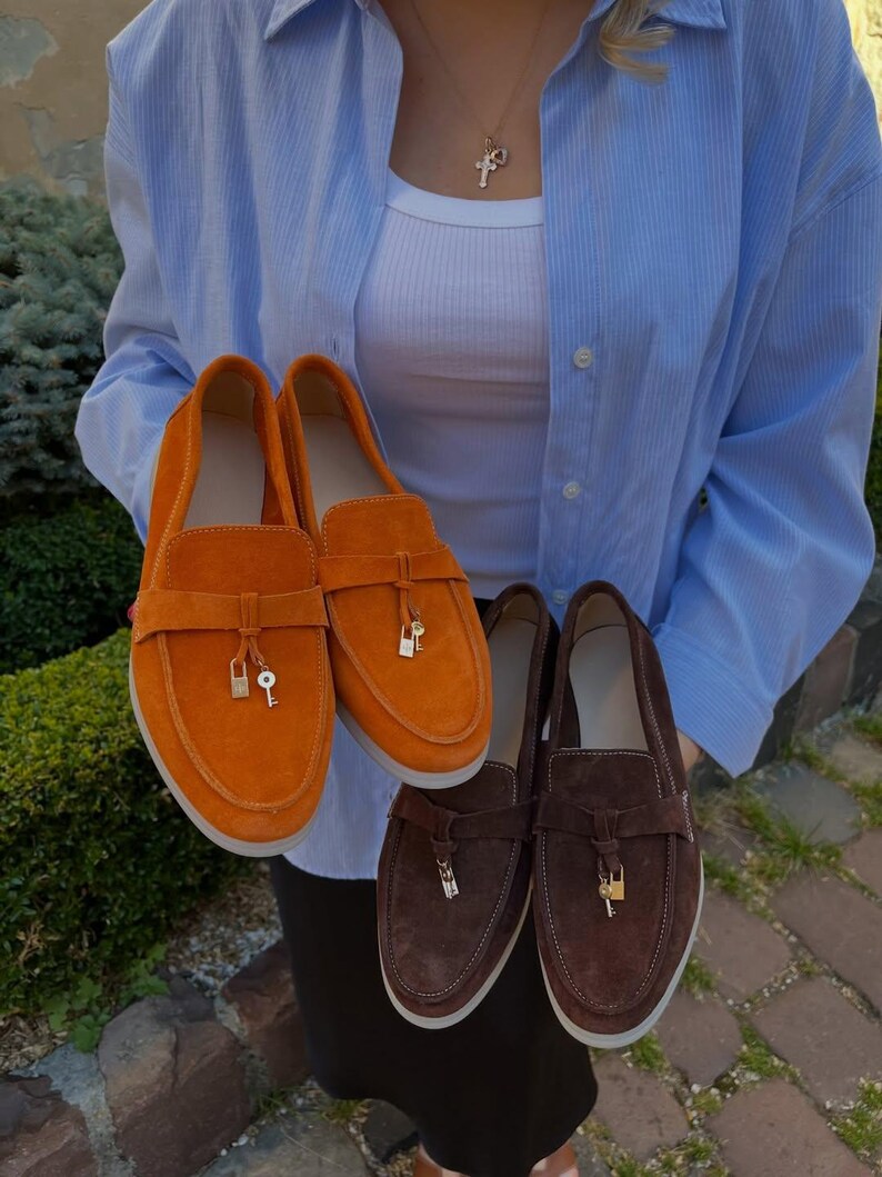 May include: A pair of bright orange suede loafers and a pair of dark brown suede loafers. Each shoe features a horizontal strap across the vamp adorned with a small gold-toned metal padlock and key charm hanging from the center of the strap.