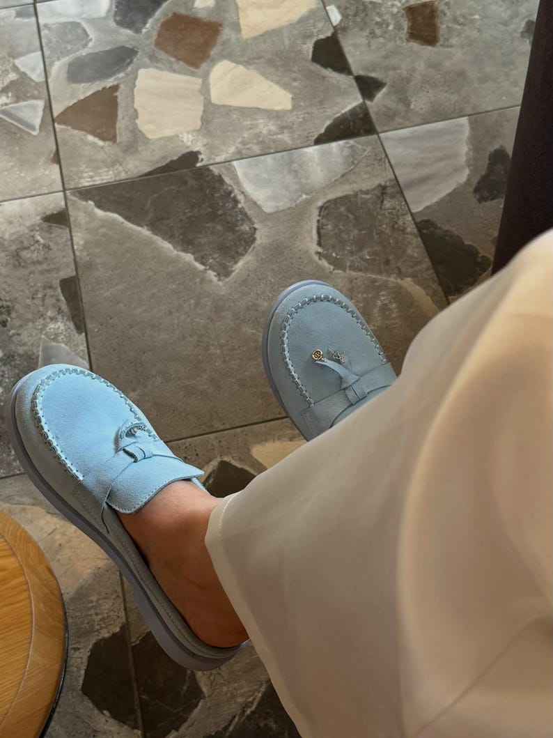 May include: Light blue suede slip-on shoes with a decorative buckle. The shoes have a rounded toe and a low heel. The image shows the shoes from a top-down perspective, with a person wearing them. The shoes are paired with cream-colored pants.