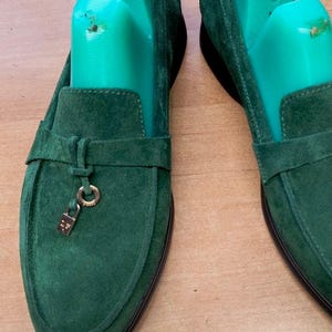 May include: A pair of dark green suede loafers featuring a horizontal strap across the vamp and a small gold-toned metal padlock charm hanging from a ring. Each shoe contains a bright green plastic shoe tree marked with the size 39.