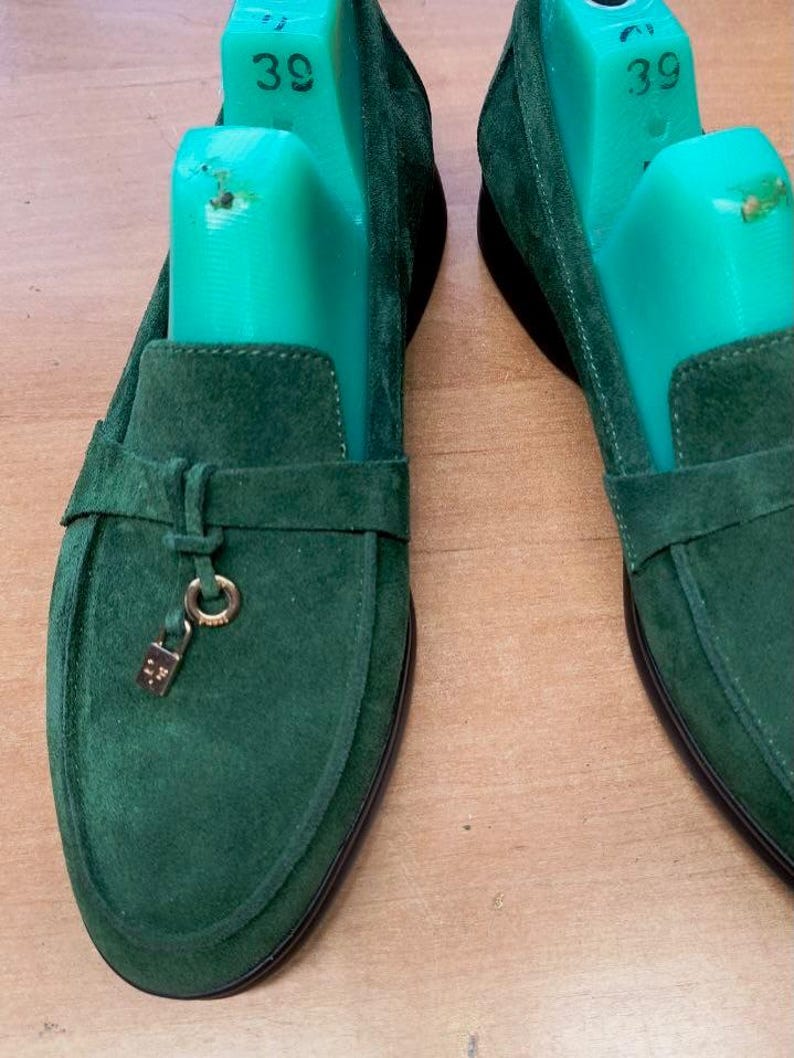 May include: A pair of dark green suede loafers featuring a horizontal strap across the vamp and a small gold-toned metal padlock charm hanging from a ring. Each shoe contains a bright green plastic shoe tree marked with the size 39.