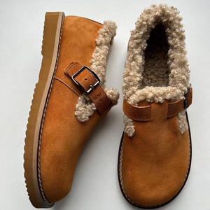 Könnte beinhalten: Ein Paar braune Wildleder-Ankle Boots mit Shearling-Futter. Die Stiefel haben einen Schnallenverschluss und eine dicke, hellbraune Sohle. Der Shearling-Besatz ist hellbeige und das Wildleder ist ein warmes, erdiges Braun.