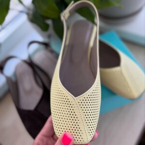 May include: Pale yellow perforated leather slingback flats with a square toe. The shoes have a thin ankle strap and a neutral-colored insole. A second pair of brown shoes is visible in the background.