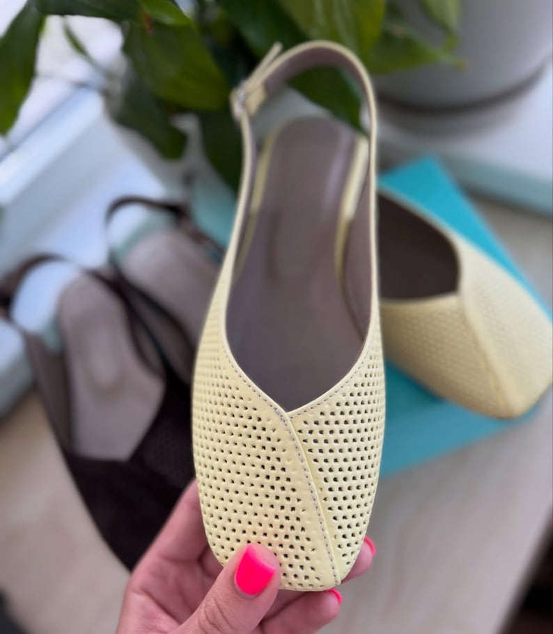 May include: Pale yellow perforated leather slingback flats with a square toe. The shoes have a thin ankle strap and a neutral-colored insole. A second pair of brown shoes is visible in the background.