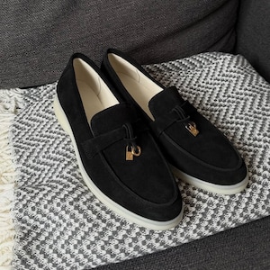 May include: A pair of black suede loafers featuring a gold-toned metal padlock charm attached to a decorative strap across the vamp. The shoes have a smooth cream-colored interior lining and a thick, translucent white rubber sole for traction.