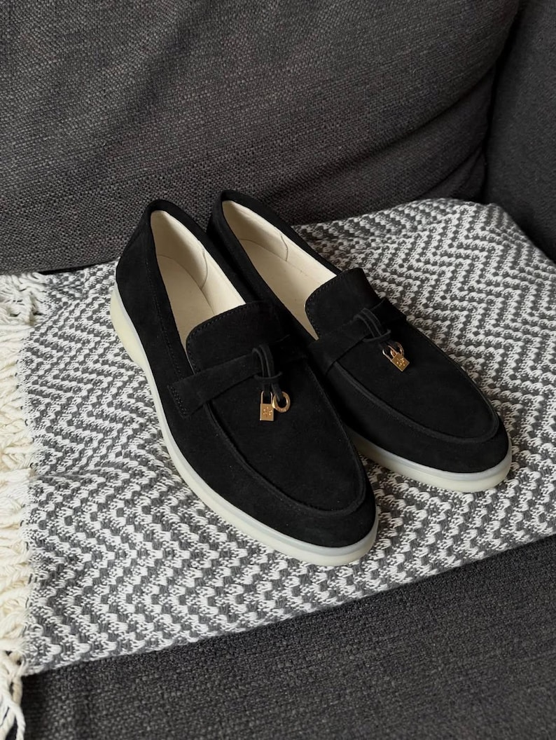 May include: A pair of black suede loafers featuring a gold-toned metal padlock charm attached to a decorative strap across the vamp. The shoes have a smooth cream-colored interior lining and a thick, translucent white rubber sole for traction.