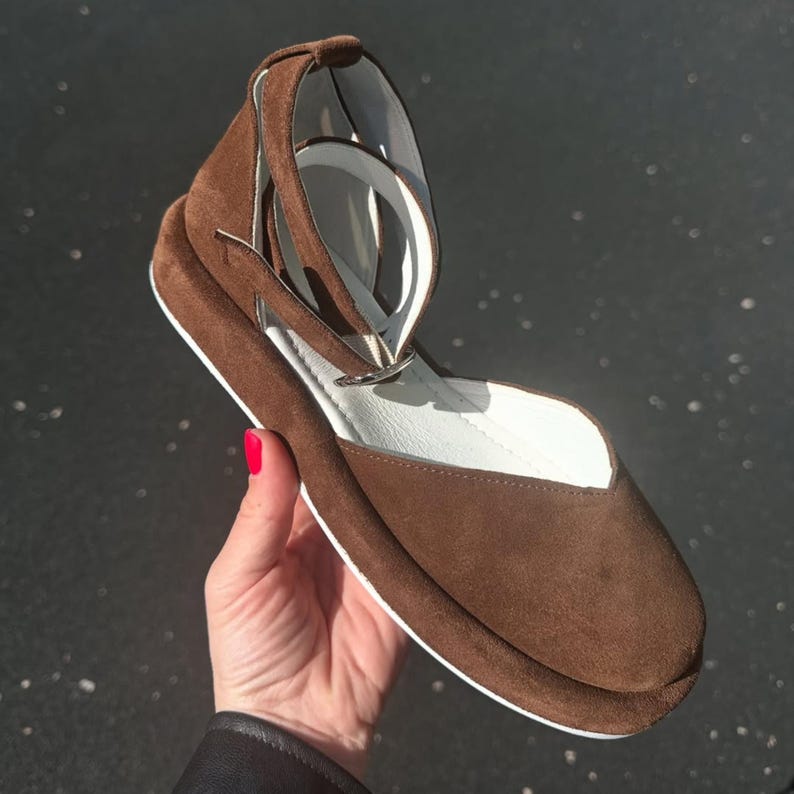 May include: A brown suede shoe with a white sole and ankle strap. The shoe has a rounded toe and a small heel. The ankle strap is secured with a silver buckle. The interior of the shoe is white.