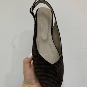 May include: A dark brown slingback flat shoe with a square toe. The shoe has a textured surface and a thin strap that buckles at the ankle. The interior is a light beige color.