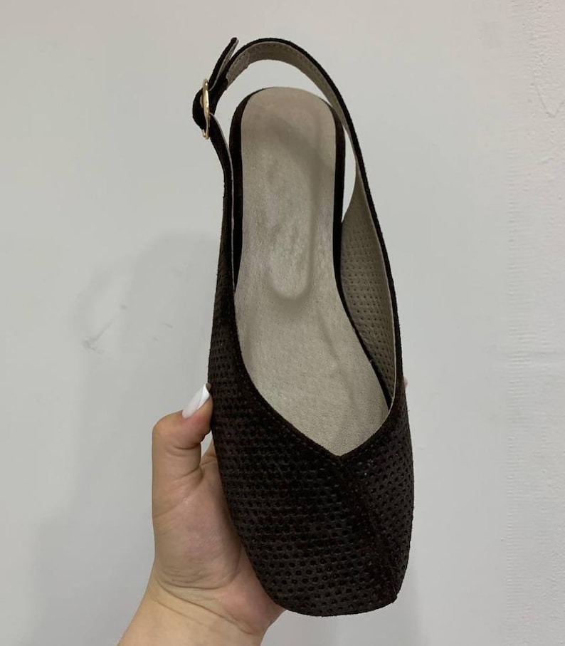 May include: A dark brown slingback flat shoe with a square toe. The shoe has a textured surface and a thin strap that buckles at the ankle. The interior is a light beige color.