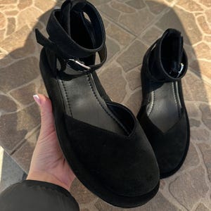 May include: A pair of black suede flats with ankle straps. The shoes have a rounded toe and a small heel. The ankle straps fasten with a silver buckle. The shoes are displayed on a stone surface.
