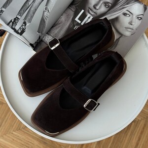 May include: A pair of dark brown Mary Jane style flats with square toes and silver-tone buckles. The shoes have a strap across the top and a brown sole. The shoes are displayed on a white surface with a magazine in the background.