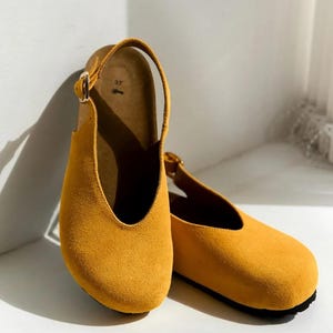 May include: A pair of mustard yellow suede slingback clogs with a rounded toe and a gold-toned metal buckle on the adjustable heel strap. The interior footbed is tan and features the number 37 printed in black near the heel area.