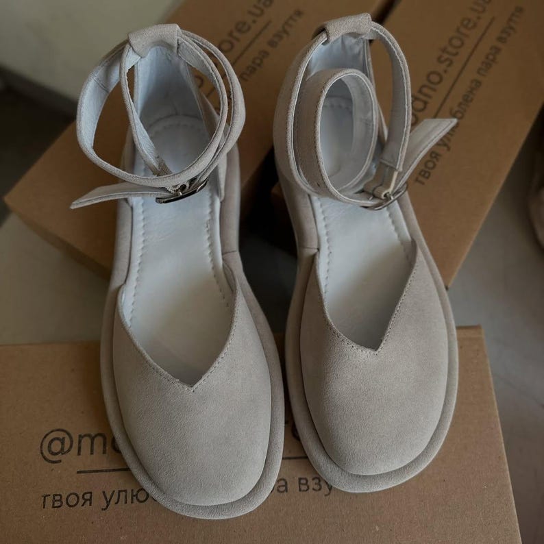 May include: A pair of light beige suede sandals with ankle straps and silver-tone buckles. The sandals have a rounded toe and a V-shaped vamp. The soles are a slightly darker shade of beige. The sandals are displayed on a cardboard box.