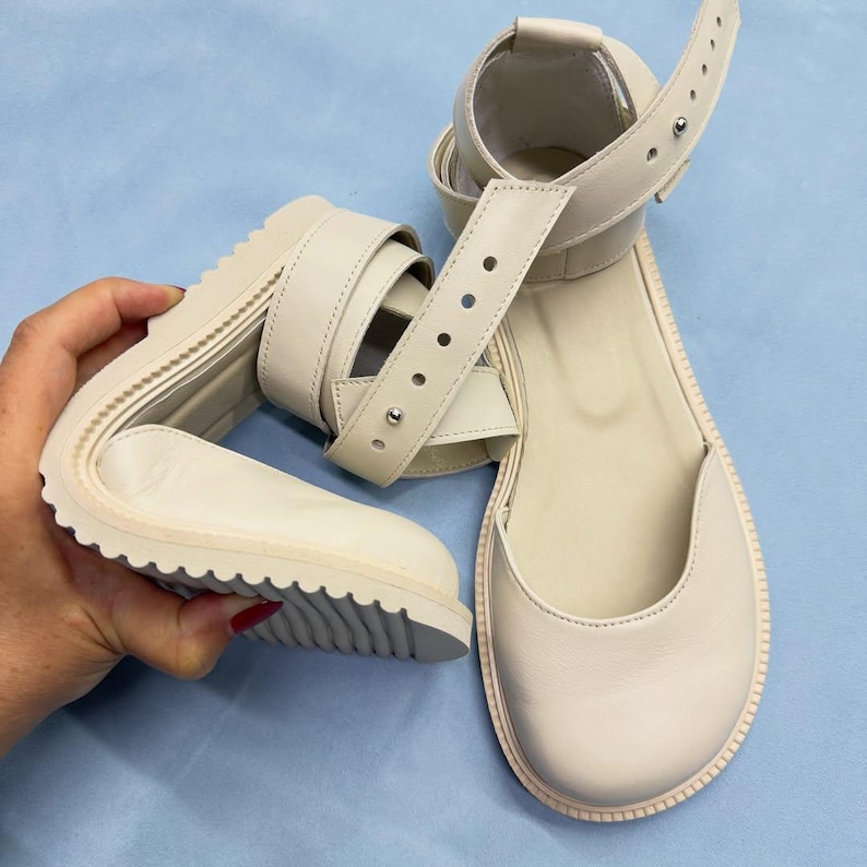May include: Off-white leather sandals with a thick, ridged sole. The sandals feature an ankle strap with multiple adjustment holes and a buckle closure. The design includes a rounded toe and a strap that wraps around the foot.