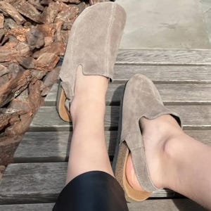 Può includere: Un paio di zoccoli in camoscio color talpa con design a cinturino. Gli zoccoli hanno un plantare in sughero e una punta arrotondata. Le scarpe sono mostrate su una panca di legno.