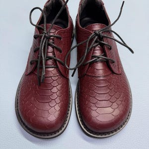 May include: A pair of burgundy leather shoes with a textured, scale-like pattern. The shoes have black laces and a rounded toe. The soles are white with a black stripe. The shoes are a classic style.