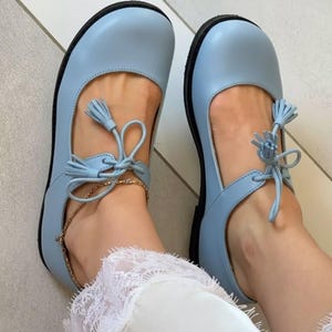 May include: Light blue Mary Jane style shoes with black soles. The shoes feature a rounded toe, a strap with a tassel, and a gold chain ankle bracelet. The shoes are made of a smooth material.