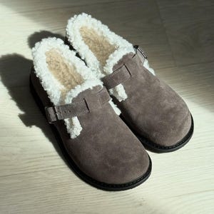 May include: A pair of gray suede clogs with a white, fluffy interior lining. The clogs have a strap with a silver buckle and a black sole. The shoes are designed for comfort and warmth.