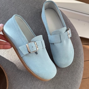 May include: A pair of light blue suede loafers with a buckle closure. The shoes have a rounded toe and a brown sole. The loafers are displayed on a gray surface.