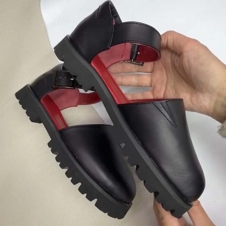 May include: Black leather sandals with a red interior and a thick, lug sole. The sandals have a strap with a buckle closure and a closed-toe design. The shoes are held up by a person's hands.