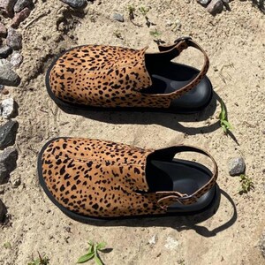 May include: A pair of leopard print sandals with black soles and adjustable ankle straps. The sandals are made of a tan material with black spots, and are set on a sandy surface with small rocks and greenery.