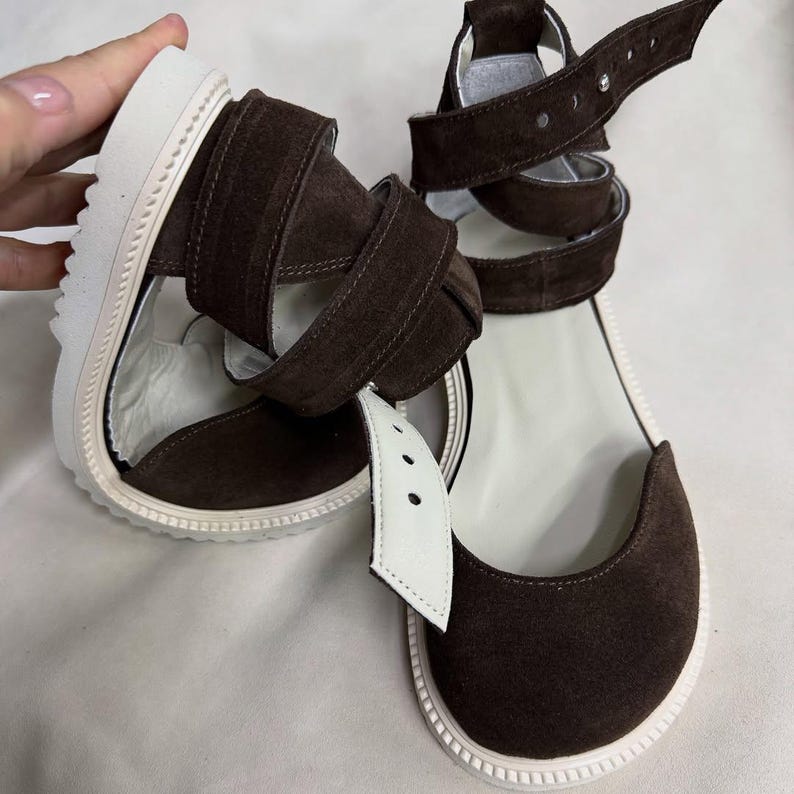 May include: Brown suede sandals with white soles and straps. The sandals feature a wrap-around ankle strap design and a white leather strap with three holes. The soles are thick and white with a textured edge.