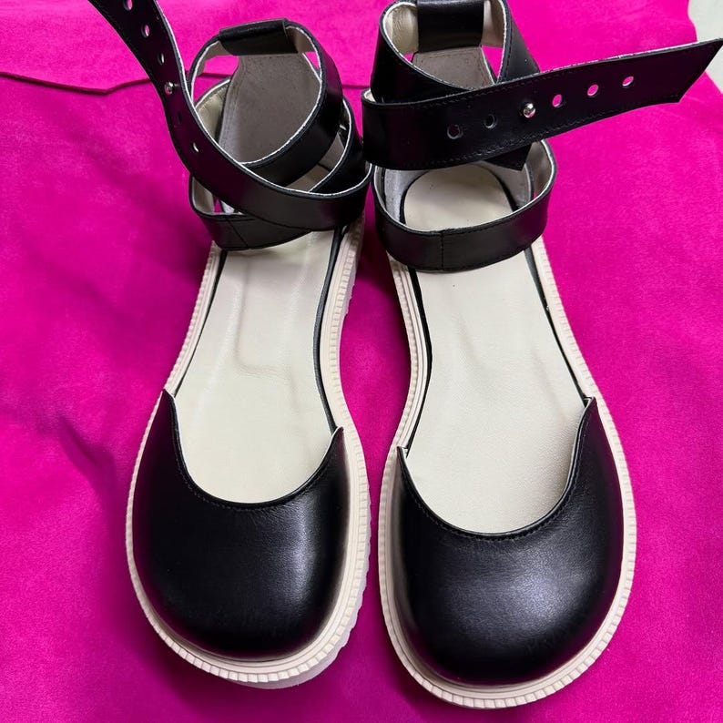 May include: A pair of black leather sandals with white soles and ankle straps. The sandals have a rounded toe design and adjustable straps. The background is a bright pink color.