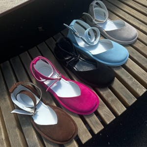 May include: A collection of colorful, flat-soled shoes with ankle straps. The shoes are in shades of brown, fuchsia, black, light blue, and gray. Each shoe has a rounded toe and a strap that buckles around the ankle. The shoes are arranged on a wooden surface.