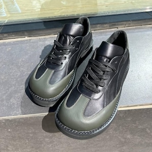 Puede incluir: Un par de zapatillas de cuero negro con punteras y suelas verde oliva. Los zapatos tienen cordones negros y un diseño cosido. Las zapatillas se exhiben sobre una superficie gris.