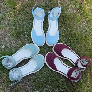 May include: A collection of flat sandals in various colors, including light blue, teal, and burgundy. Each pair features a rounded toe, ankle strap, and a white sole. The sandals are arranged on a bed of green grass.