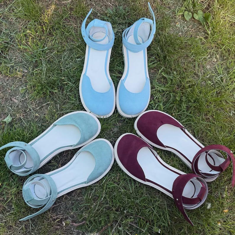 May include: A collection of flat sandals in various colors, including light blue, teal, and burgundy. Each pair features a rounded toe, ankle strap, and a white sole. The sandals are arranged on a bed of green grass.