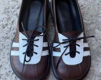 May include: A pair of brown leather shoes with white accents and black laces. The shoes have a rounded toe and a white stripe design on the front. The soles are black with a textured pattern.