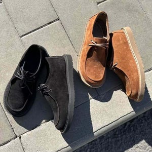 Pu&ograve; includere: Due paia di scarpe in camoscio. Un paio &egrave; nero con lacci e suole neri. L'altro paio &egrave; marrone con lacci e suole beige. Le scarpe sono esposte su una superficie di mattoni grigi.