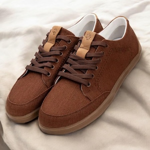 Può includere: Un paio di sneakers in tela marrone con lacci marroni e suola marrone chiaro. Le scarpe hanno l'interno bianco e un piccolo dettaglio in pelle sulla linguetta. Le sneakers sono esposte su uno sfondo di tessuto bianco.