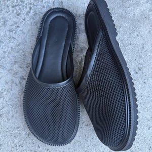 May include: A pair of black mesh slip-on sandals. The sandals have a rounded toe, a textured sole, and a mesh upper. The interior is a solid black. The sandals are designed for casual wear.