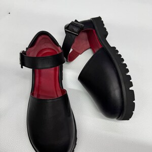 May include: Pair of black Mary Jane style shoes with a red interior. The shoes have a thick, lug sole and a buckle closure on the ankle strap. The shoes are made of a smooth material.